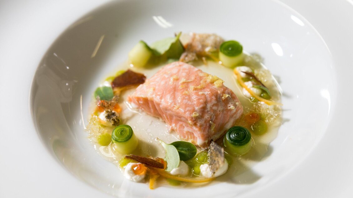 Ein zartes Stück Lachs liegt auf einem weißen Teller, umgeben von frischem Gemüse und einer klaren Sauce. Die Anrichtung wirkt elegant und einladend. | © TV Region Graz - Harry Schiffer