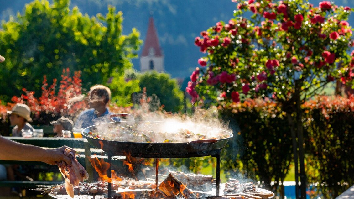 Ein gemütliches Grillfest im Freien mit rauchendem Grill und anscheinend leckerem Essen. Im Hintergrund blühen Rosen und es sind Menschen an Tischen zu sehen. | © TV Region Graz - Harry Schiffer