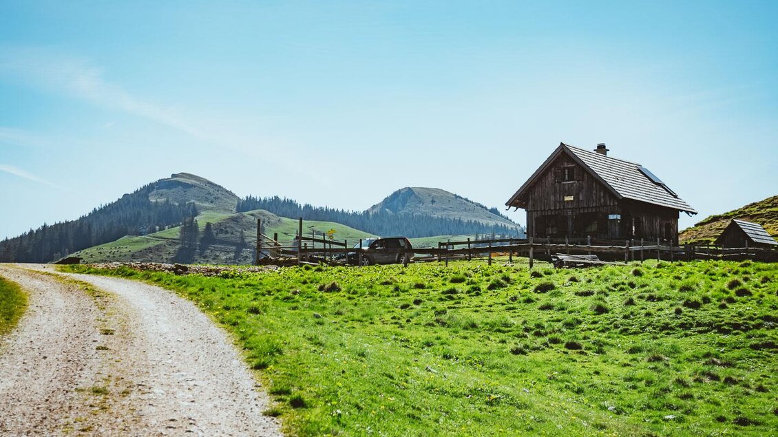 Eine malerische Landschaft mit einem traditionellen Bauernhaus und sanften Hügeln im Hintergrund. Der Weg führt durch das grüne Gras unter einem klaren blauen Himmel. | © www.mariazell.blog / Fred Lindmoser