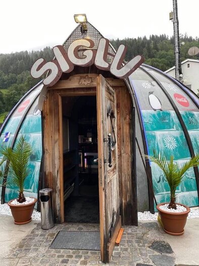 A unique, igloo-shaped building named "SIGLU". It has a wooden door and stands in front of a forested backdrop. | © Siglu-Bar Schladming