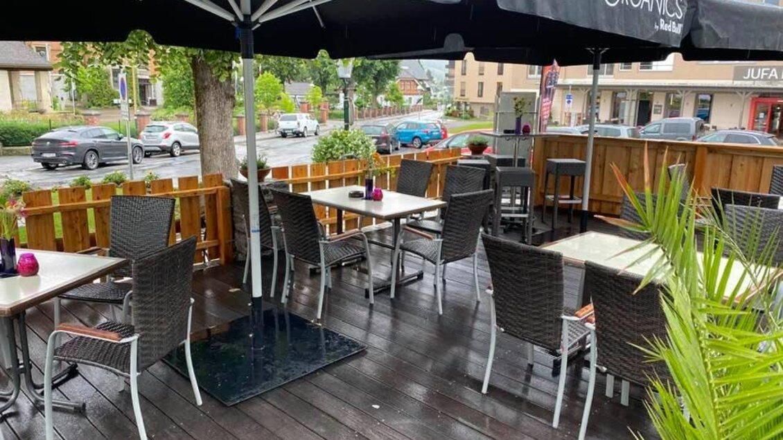 Eine gemütliche Außenterrasse eines Restaurants mit Stühlen und Tischen unter einem großen Sonnenschirm. Die Umgebung ist begrünt und einladend. | © Siglu-Bar Schladming