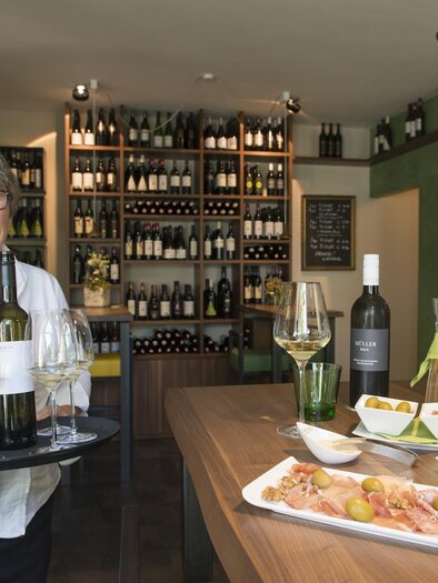 A smiling woman serves wine and snacks in a cozy wine bar. In the background, shelves with wine bottles are visible. | © Tourismusverband Feldbach/ B. Bergmann