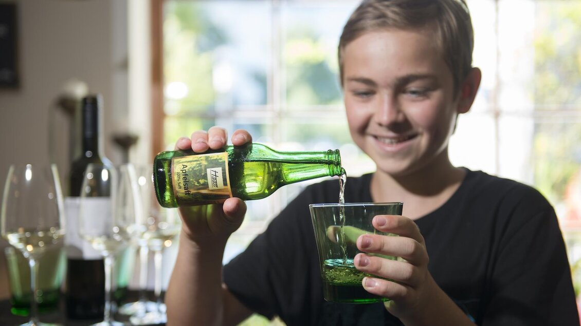 Ein Junge gießt ein Getränk aus einer grünen Flasche in ein Glas. Im Hintergrund sind Weingläser und eine Flasche Wein sichtbar. | © Sissi´s Weinbar