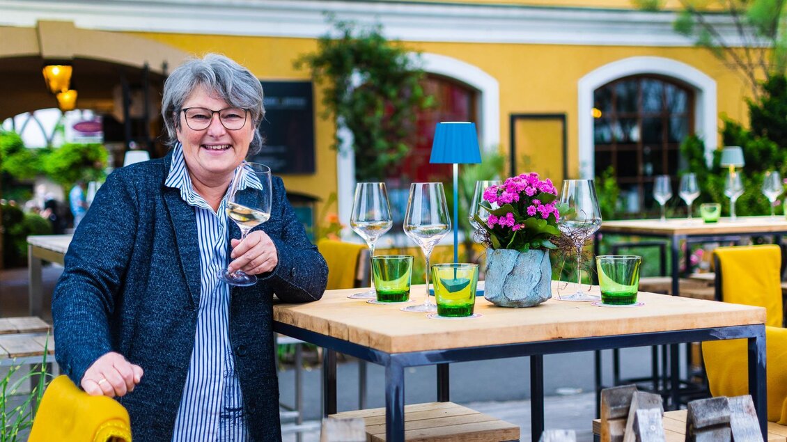 Eine Frau lächelt an einem Tisch im Freien, umgeben von Gläsern und einer Blume. Der Hintergrund zeigt ein einladendes Restaurant mit gelber Wandfarbe. | © Sissi´s Weinbar