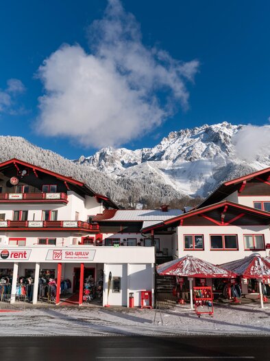 A picturesque mountain village with snow-covered mountains in the background. In the foreground, a few charming houses with red accents can be seen. | © Ski Willy