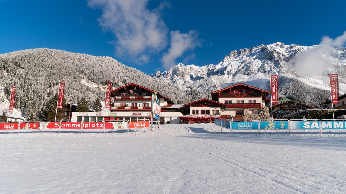 Eine verschneite Landschaft mit einem Gebäude im Vordergrund und majestätischen Bergen im Hintergrund. Der Himmel ist klar und blau, was eine winterliche Atmosphäre schafft. | © SkiWilly