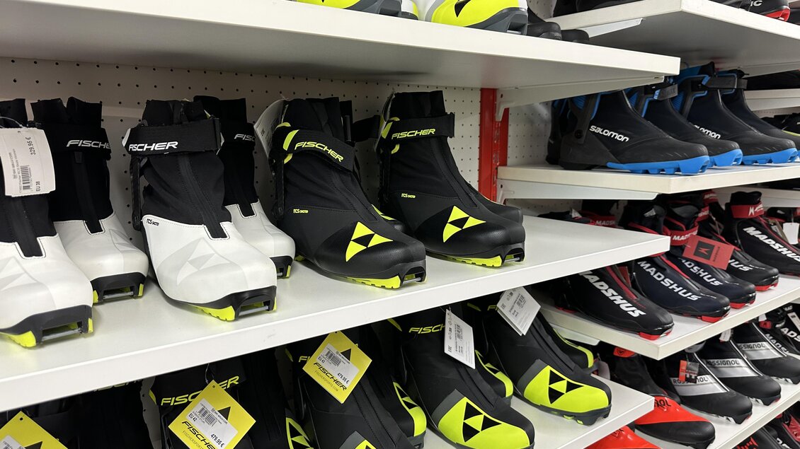 Skischuhe in verschiedenen Farben und Designs sind auf Regalen ausgestellt. Die Schuhe sind hauptsächlich schwarz mit auffälligen gelben und blauen Akzenten. | © SkiWilly