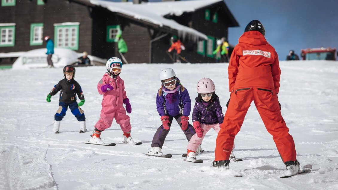 Eine Gruppe von Kindern in farbigen Skianzügen übt das Skifahren auf einer verschneiten Piste. Im Hintergrund ist ein Holzhaus zu sehen. | © RUDY'SHCOOL