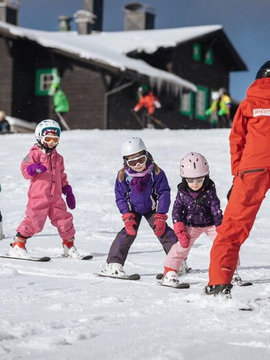 A group of children in colorful ski suits are practicing skiing on a snowy slope. In the background, a wooden house can be seen. | © RUDY'SHCOOL