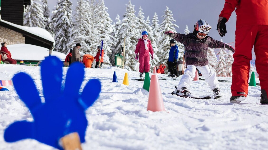 Kinder lernen das Skifahren auf einem verschneiten Hang. Im Hintergrund sind Erwachsene und bunte Pylonen zu sehen. | © RUDY'SHCOOL