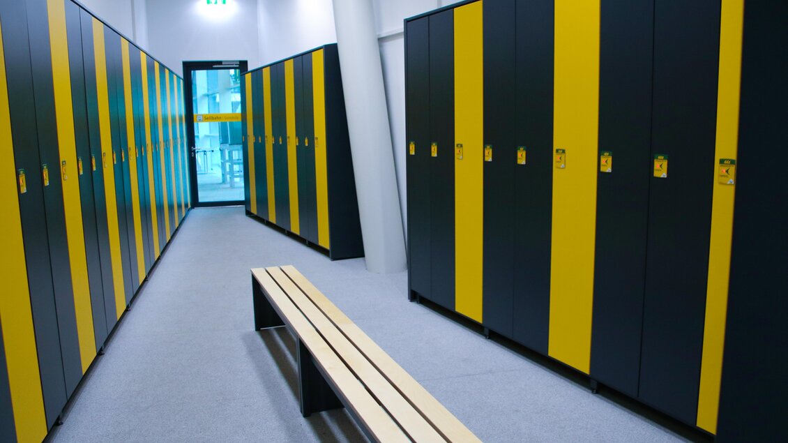 Ein moderner Umkleideraum mit schwarzen und gelben Spinden. In der Mitte steht eine Holzbank und ein Fenster führt nach außen. | © Planai