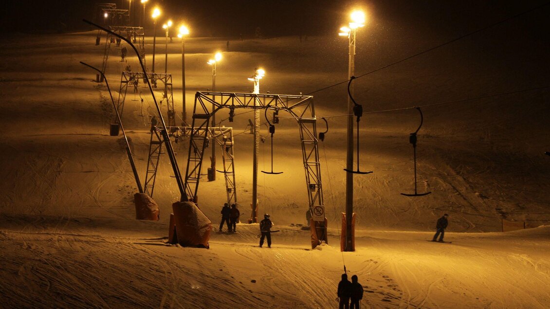 Eine verschneite Piste bei Nacht mit beleuchteten Skiliften. Einige Leute sind auf der Piste zu sehen. | © Zlaim Lift