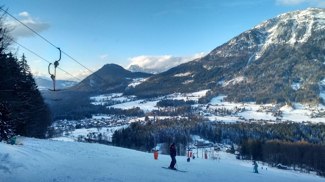 Eine malerische Berglandschaft mit verschneiten Hängen und einem klaren Himmel. Skifahrer genießen die Piste in der Nähe eines kleinen Dorfes. | © Zlaim Lift/Franz Steinegger