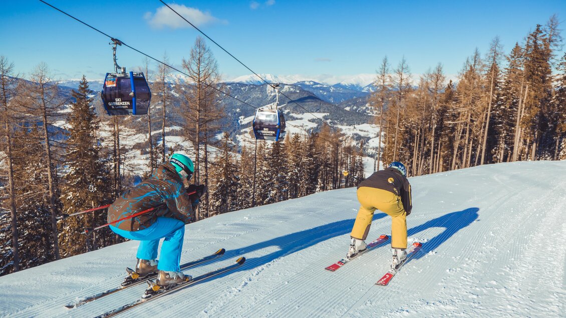 Zwei Skifahrer stehen auf einer verschneiten Piste mit Bäumen im Hintergrund. Im Himmel sind Gondeln zu sehen, die zu den Bergen fahren. | © Tourismusverband Murau