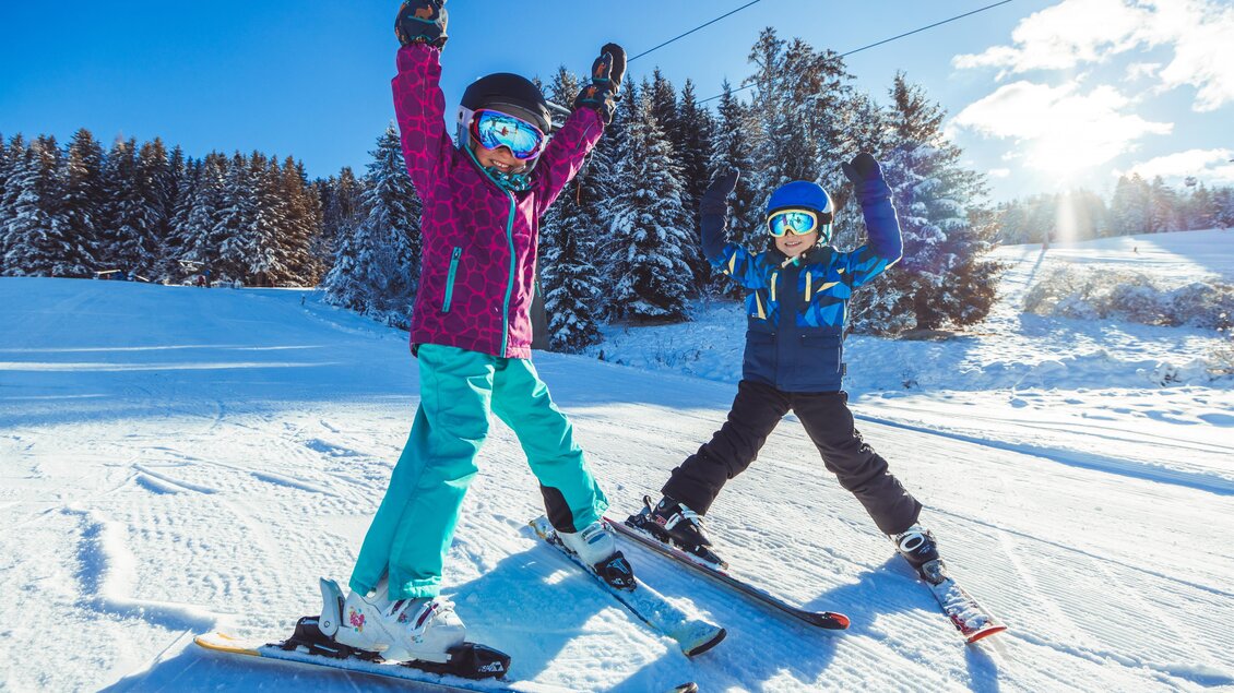 Zwei Kinder stehen auf Skiern in einer verschneiten Landschaft und jubeln fröhlich. Im Hintergrund sind esige Bäume und ein blauer Himmel zu sehen. | © Tourismusverband Murau