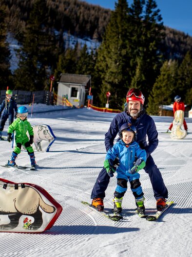 A cheerful father and his son are skiing together in a sunny ski resort. In the background, other children and ski lodges can be seen. | © Lachtal-Lifte u. Seilbahnen GmbH