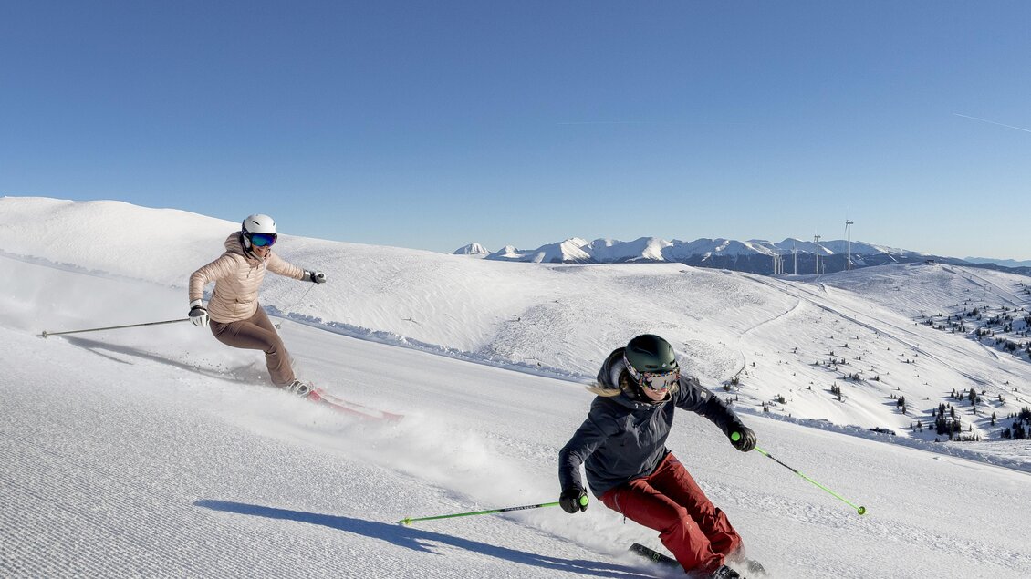 Zwei Skifahrer fahren auf einer schneebedeckten Piste. Im Hintergrund sind Berge und ein klarer blauer Himmel zu sehen. | © Lachtal-Lifte u. Seilbahnen GmbH