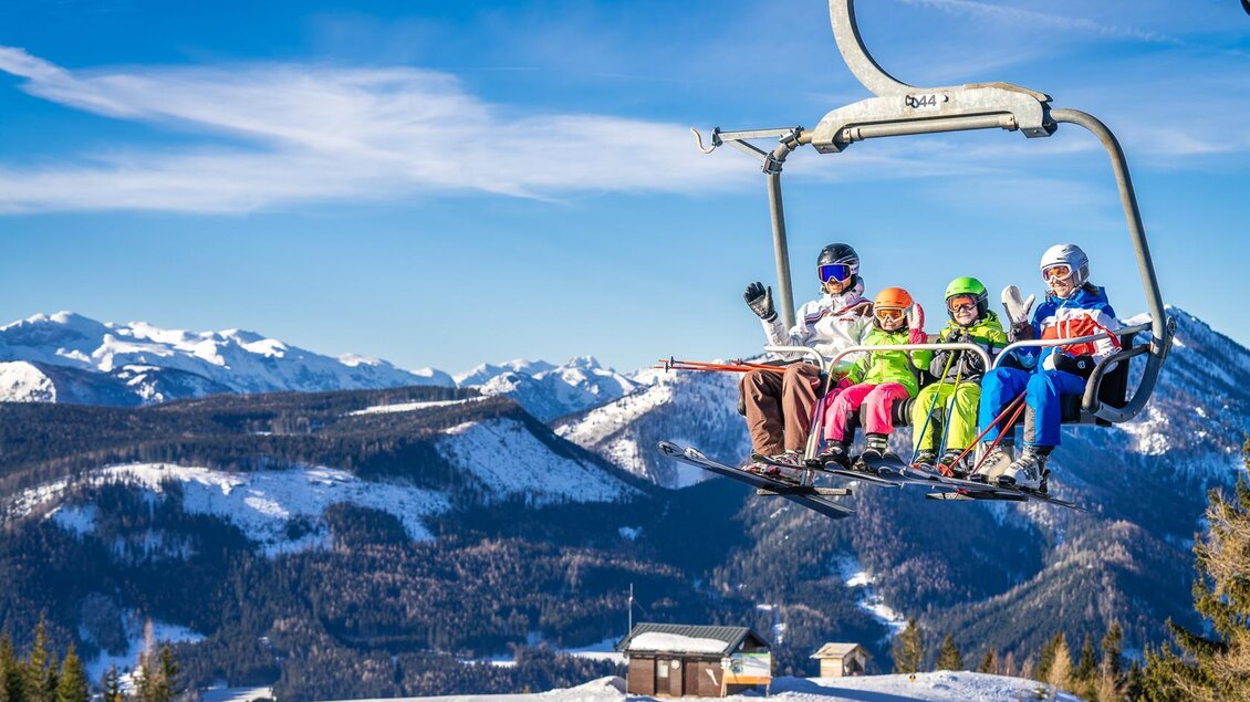 Eine Gruppe von Skifahrern fährt in einem Sessellift über schneebedeckte Berge. Der Himmel ist klar und sonnig. | © Mariazeller Bürgeralpe | Rudy Dellinger