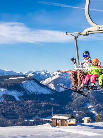 A group of skiers is riding a chairlift over snow-covered mountains. The sky is clear and sunny. | © Mariazeller Bürgeralpe | Rudy Dellinger