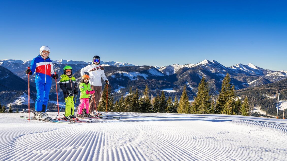 Eine Familie steht auf einer schneebedeckten Piste mit Skiausrüstung. Im Hintergrund sind majestätische Berge und ein klarer blauer Himmel zu sehen. | © Mariazeller Bürgeralpe | Rudy Dellinger
