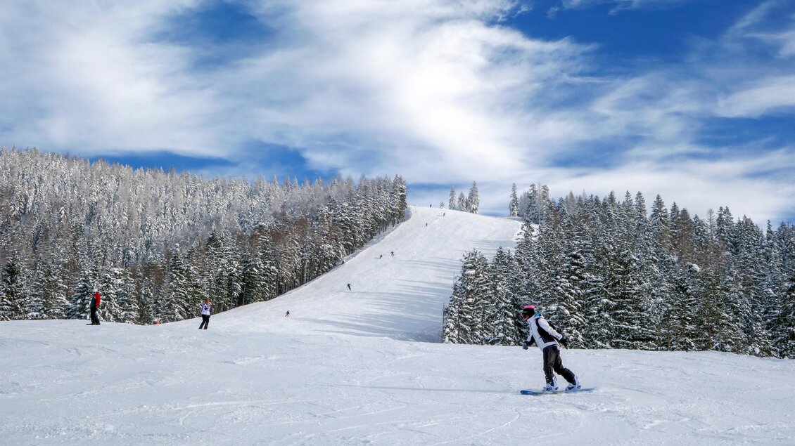 Eine schneebedeckte Landschaft mit einem Skihang und vielen Bäumen im Hintergrund. Einige Menschen sind beim Snowboarden und Skifahren zu sehen. | © TV Hochsteiermark | Fred Lindmoser