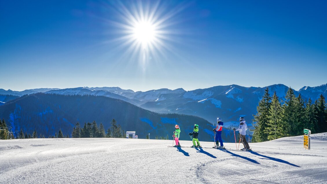 Eine Gruppe von Skifahrern genießt einen sonnigen Tag auf der Piste. Im Hintergrund sind schneebedeckte Berge und ein blauer Himmel zu sehen. | © Mariazeller Bürgeralpe | Rudy Dellinger