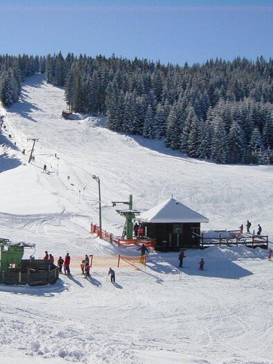 A picturesque ski slope with freshly fallen snow and people skiing. In the background, there are dense, snow-covered trees and a mountain station. | © Schigebiet Modriach-Winkel