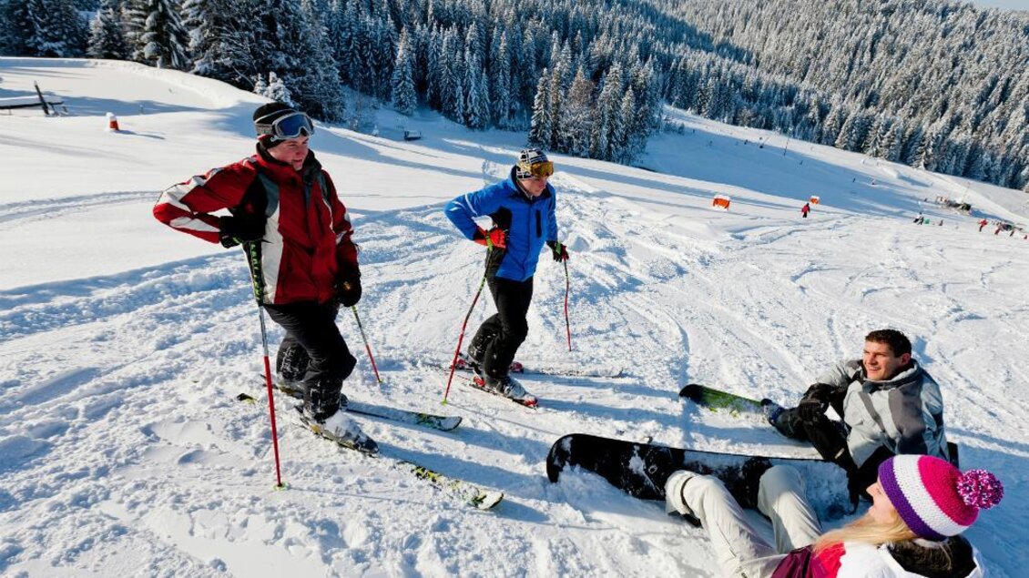 Eine Gruppe von Skifahrern entspannt sich auf einer schneebedeckten Piste. Im Hintergrund sind verschneite Bäume und Berge zu sehen. | © Ikarus | Tom Lamm