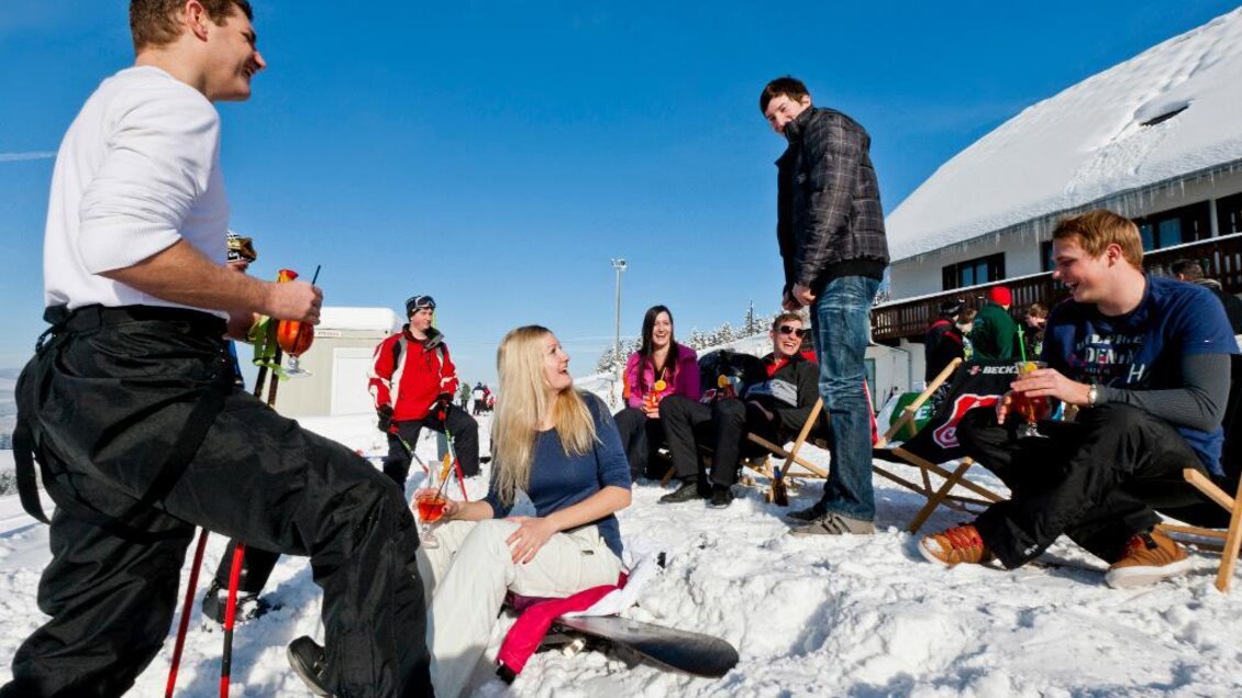 Eine Gruppe von Menschen genießt die Zeit im Schnee. Einige sitzen auf Liegen, während andere sich unterhalten und Ski-Equipment halten. | © Ikarus | Tom Lamm