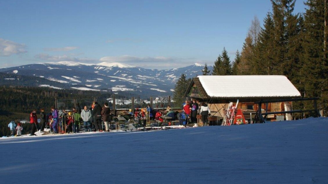 Ein wintersportlicher Ausblick mit Menschen, die sich in der Nähe einer Skihütte aufhalten. Die Umgebung ist schneebedeckt und von Bergen und Bäumen umgeben. | © Steirische Rucksackdörfer