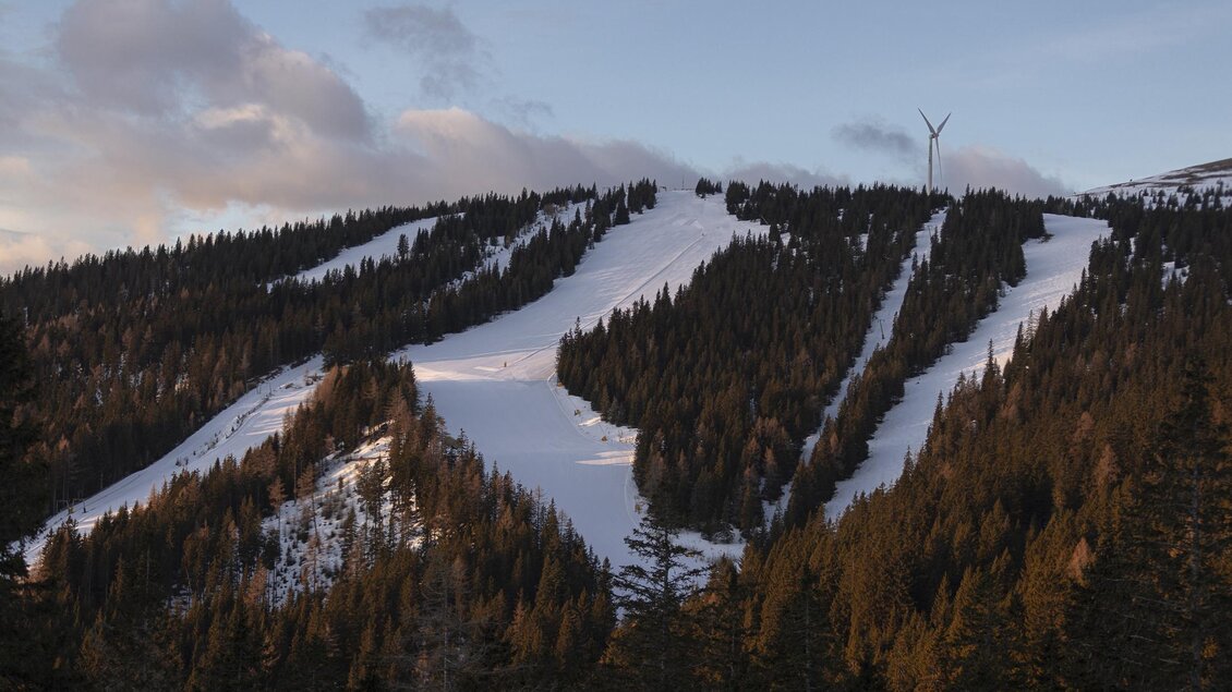 Eine verschneite Berglandschaft mit Skipisten und hohen Tannen. Im Hintergrund sind einige Windkraftanlagen sichtbar. | © Salzstiegl - Nicole Seiser