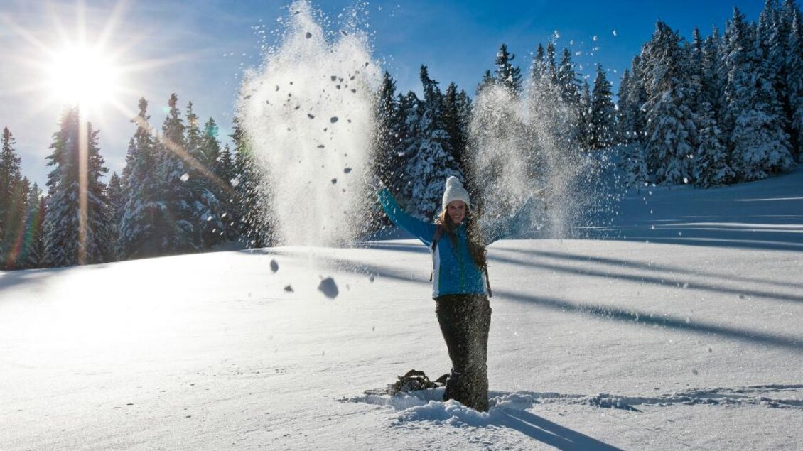 Eine Person steht im Schnee und wirbelt Schneekristalle in die Luft. Im Hintergrund sind schneebedeckte Bäume und ein blauer Himmel zu sehen. | © Ikarus | Tom Lamm