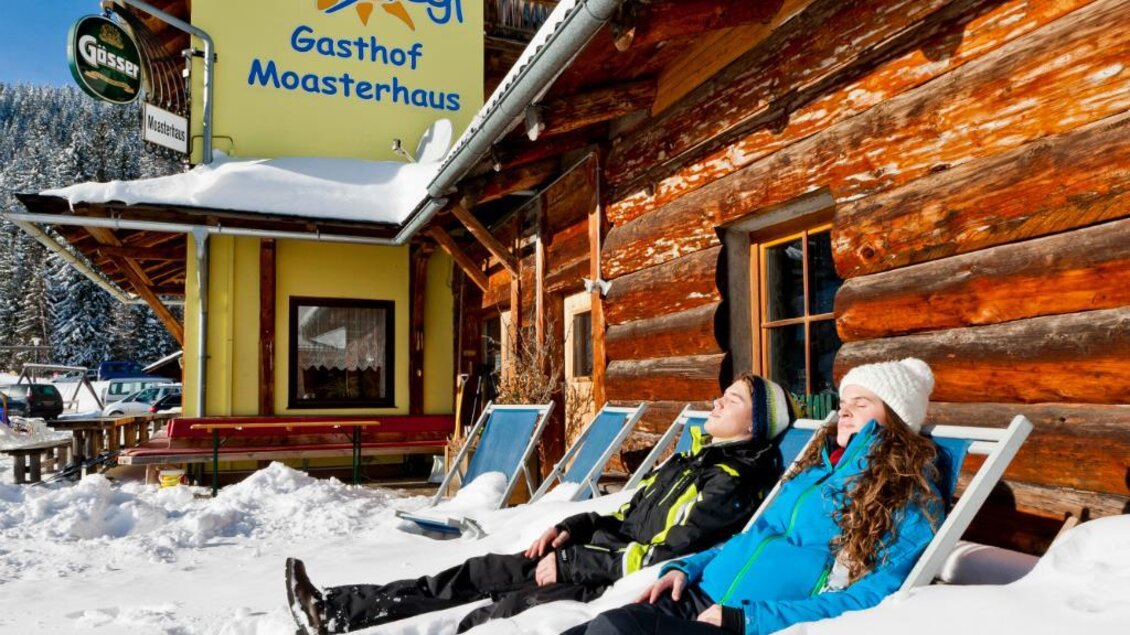 Zwei Personen entspannen im Schnee vor einem Gasthaus. Im Hintergrund ist ein Holzgebäude mit dem Schild "Gasthof Moasterhaus" zu sehen. | © Ikarus | Tom Lamm