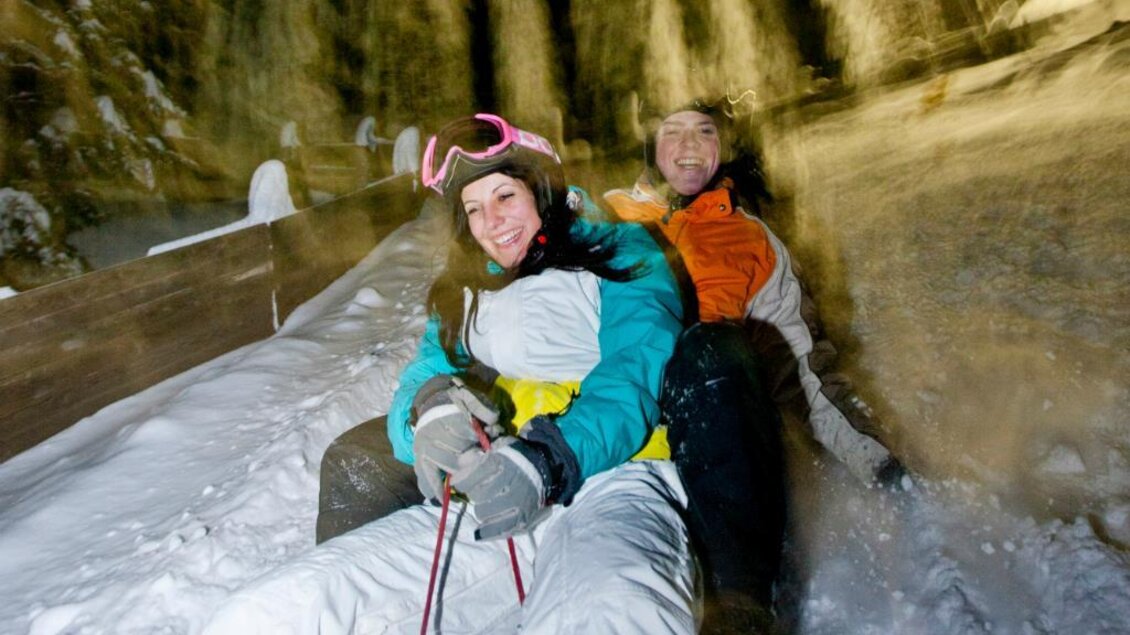 Zwei fröhliche Menschen fahren im Schnee auf einem Schlitten. Es ist eine Nachtaufnahme mit verschneiten Bäumen im Hintergrund. | © Ikarus | Tom Lamm