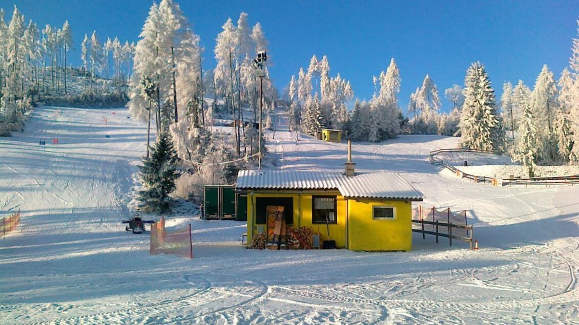Eine verschneite Winterlandschaft mit einem gelben Chalet. Im Hintergrund sind schneebedeckte Bäume und eine Skipiste zu sehen. | © Rudolf Sabetz