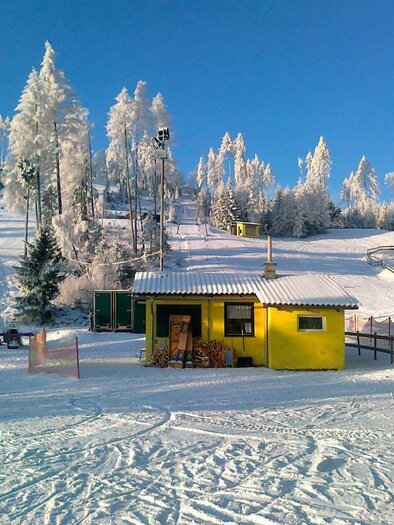 Eine verschneite Winterlandschaft mit einem gelben Chalet. Im Hintergrund sind schneebedeckte Bäume und eine Skipiste zu sehen. | © Rudolf Sabetz