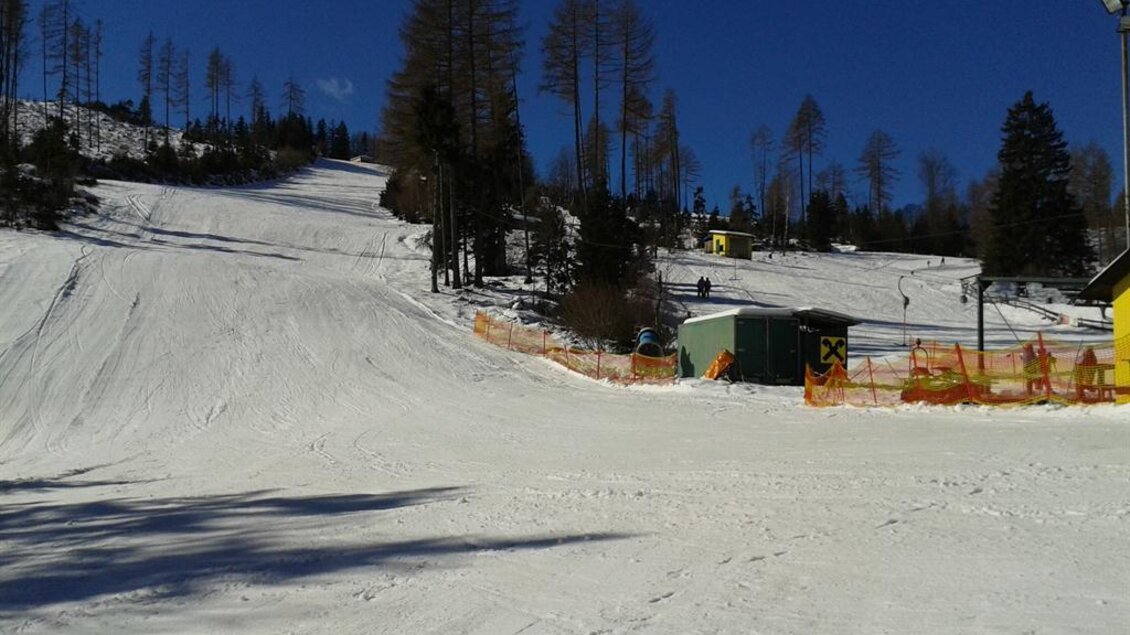 Eine schneebedeckte Piste mit einem klaren blauen Himmel. Im Hintergrund sind Bäume und eine kleine Skihütte zu sehen. | © Rudolf Sabetz
