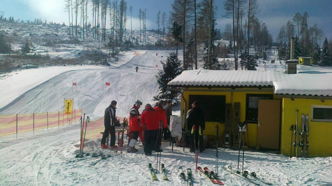 Ein Skigebiet mit einer gelben Hütte und mehreren Skifahrern. Im Hintergrund sind schneebedeckte Pisten und Bäume zu sehen. | © Rudolf Sabetz