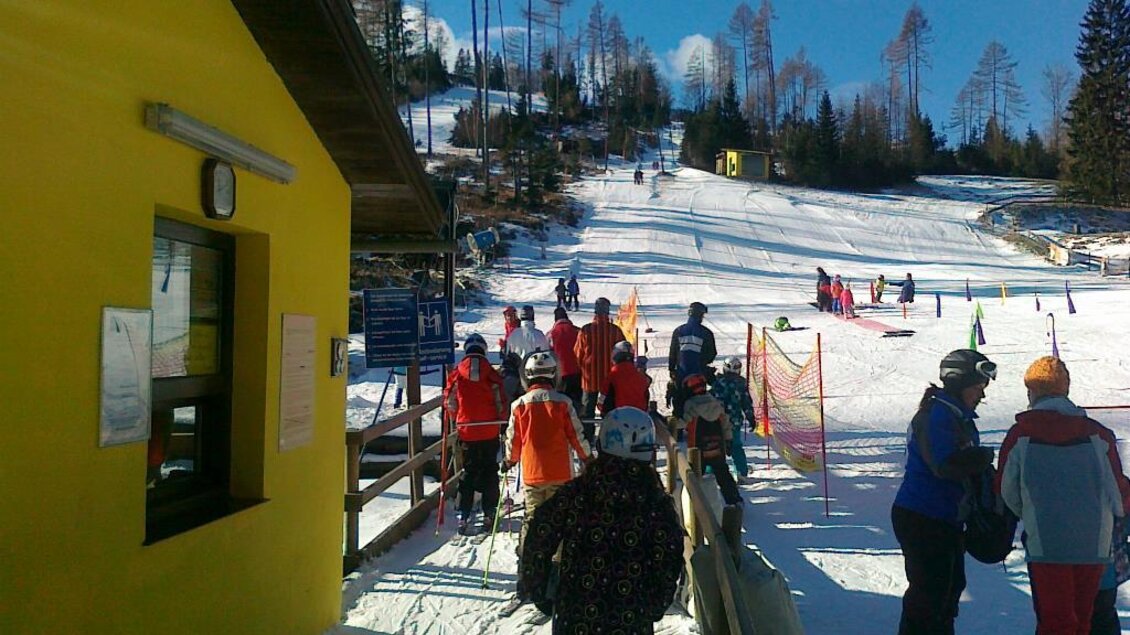 Ein belebter Skiort mit vielen Menschen, die auf die Piste gehen. Ein gelbes Gebäude ist im Vordergrund sichtbar, umgeben von schneebedeckten Hügeln. | © Rudolf Sabetz