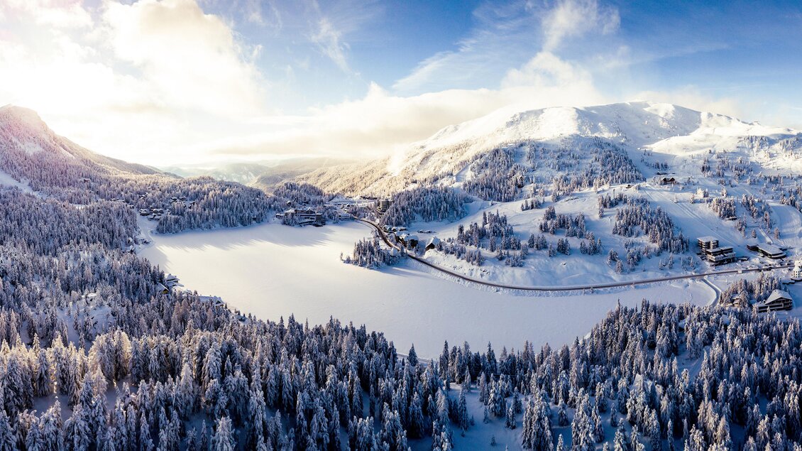Eine winterliche Landschaft mit verschneiten Bergen und einem zugefrorenen See. Dichter Nadelwald umrahmt die malerische Szenerie. | © TMG Turracher Höhe Marketing GmbH