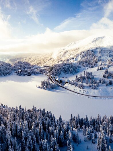 A winter landscape with snowy mountains and a frozen lake. Dense coniferous forest frames the picturesque scenery. | © TMG Turracher Höhe Marketing GmbH