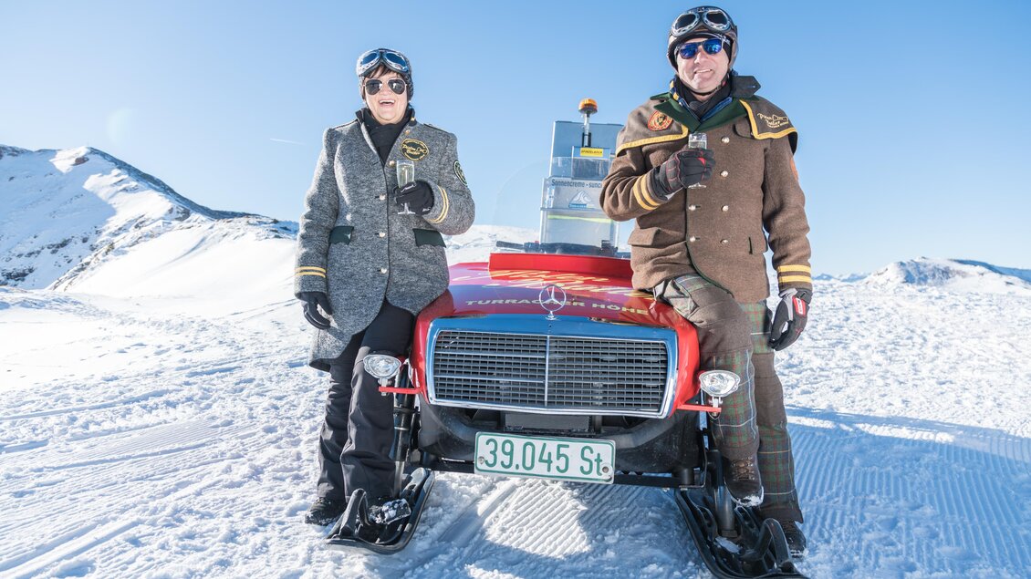 Zwei Personen in historischen Outfits stehen auf dem Schnee neben einem roten Mercedes. Im Hintergrund sind schneebedeckte Berge und ein klarer blauer Himmel zu sehen. | © TMG Turracher Höhe Marketing GmbH