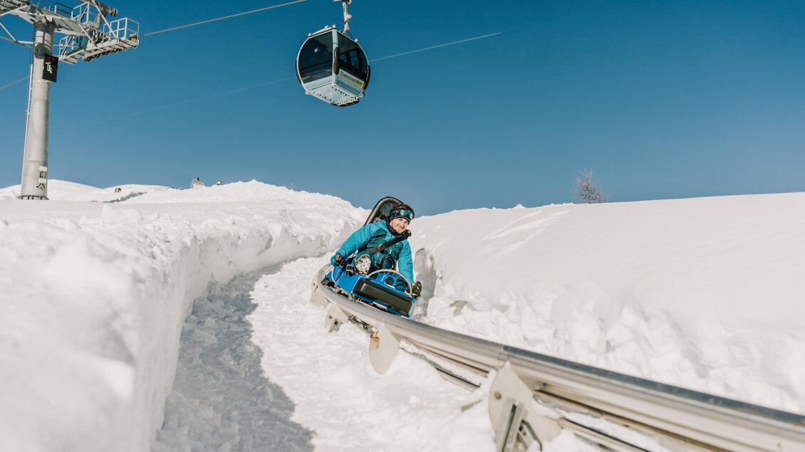 Ein Skifahrer fährt auf einer Rodelbahn durch den Schnee. Im Hintergrund sieht man eine Gondel, die in den klaren blauen Himmel schwebt. | © TMG Turracher Höhe Marketing GmbH
