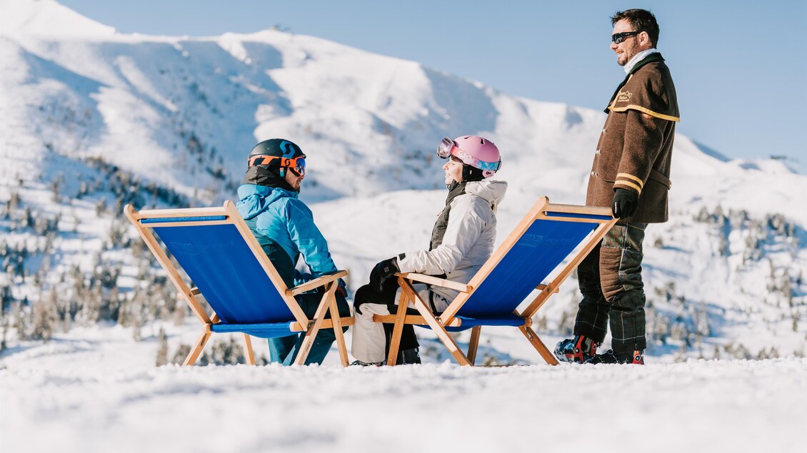 Zwei Personen entspannen auf Liegestühlen im Schnee, während ein dritter Mann steht. Im Hintergrund sind schneebedeckte Berge und blauer Himmel zu sehen. | © TMG Turracher Höhe Marketing GmbH