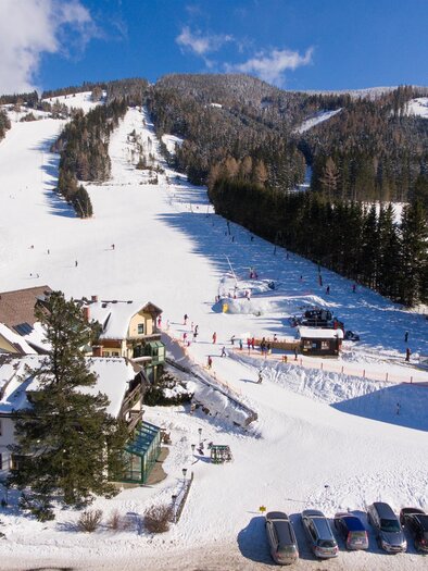 A snow-covered mountain landscape with skiers on the slopes. Below are cozy chalets and parked cars. | © Moscher