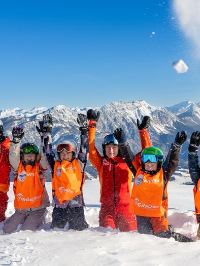 Ski instructor and children in orange vests kneel in deep snow, joyfully raising their arms up; in the background, a snowy mountain panorama under a blue sky. | © Skischule Tritscher