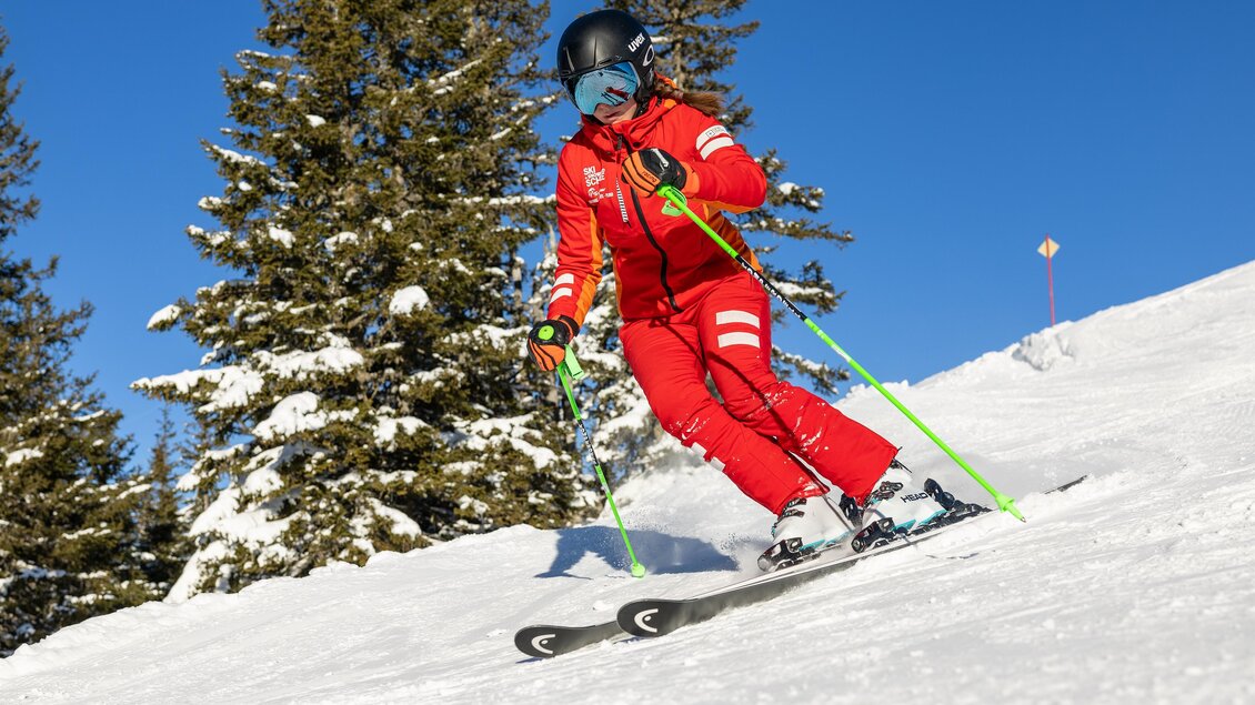 Skilehrerin fährt in rotem Skianzug eine präparierte Piste hinunter; im Hintergrund verschneite Bäume und blauer Himmel. | © Skischule Tritscher