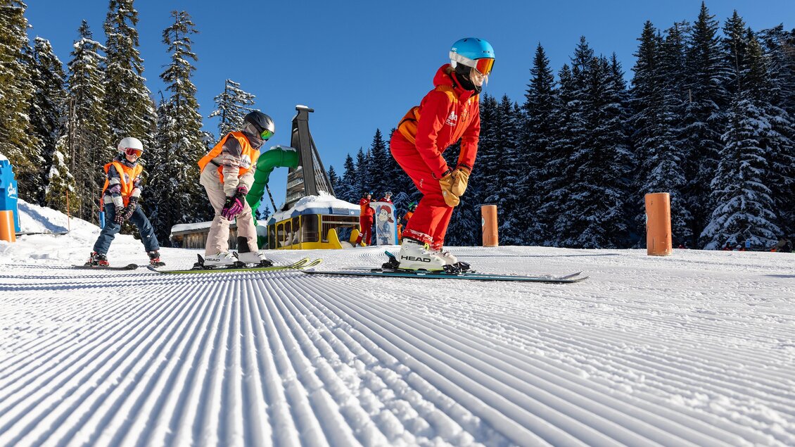 Skilehrerin fährt vorneweg, mehrere Kinder folgen ihr auf einer flachen Übungspiste; gerillter Schnee im Vordergrund, Wald im Hintergrund. | © Skischule Tritscher