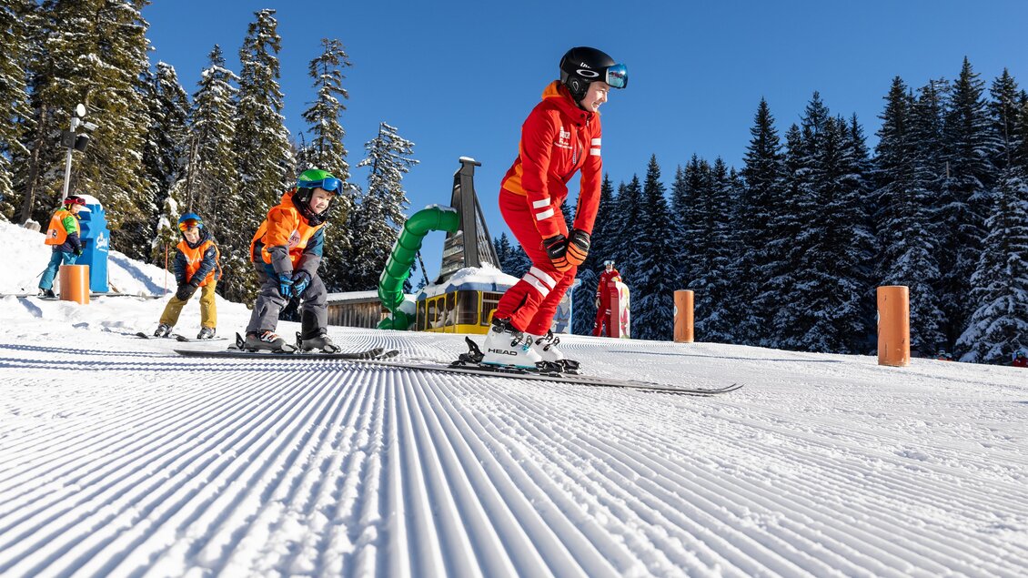 Skilehrerin und Kinder fahren gemeinsam in einer Linie über eine Übungspiste; orangefarbene Westen, verschneiter Wald und Skischulgelände im Hintergrund. | © Skischule Tritscher