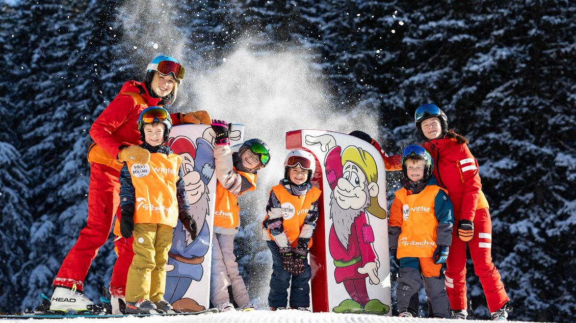 Skilehrerinnen und Kinder stehen lachend neben bunten Figuren im Schnee; aufgewirbelter Schnee sorgt für spielerische Stimmung vor dunklem Winterwald. | © Skischule Tritscher